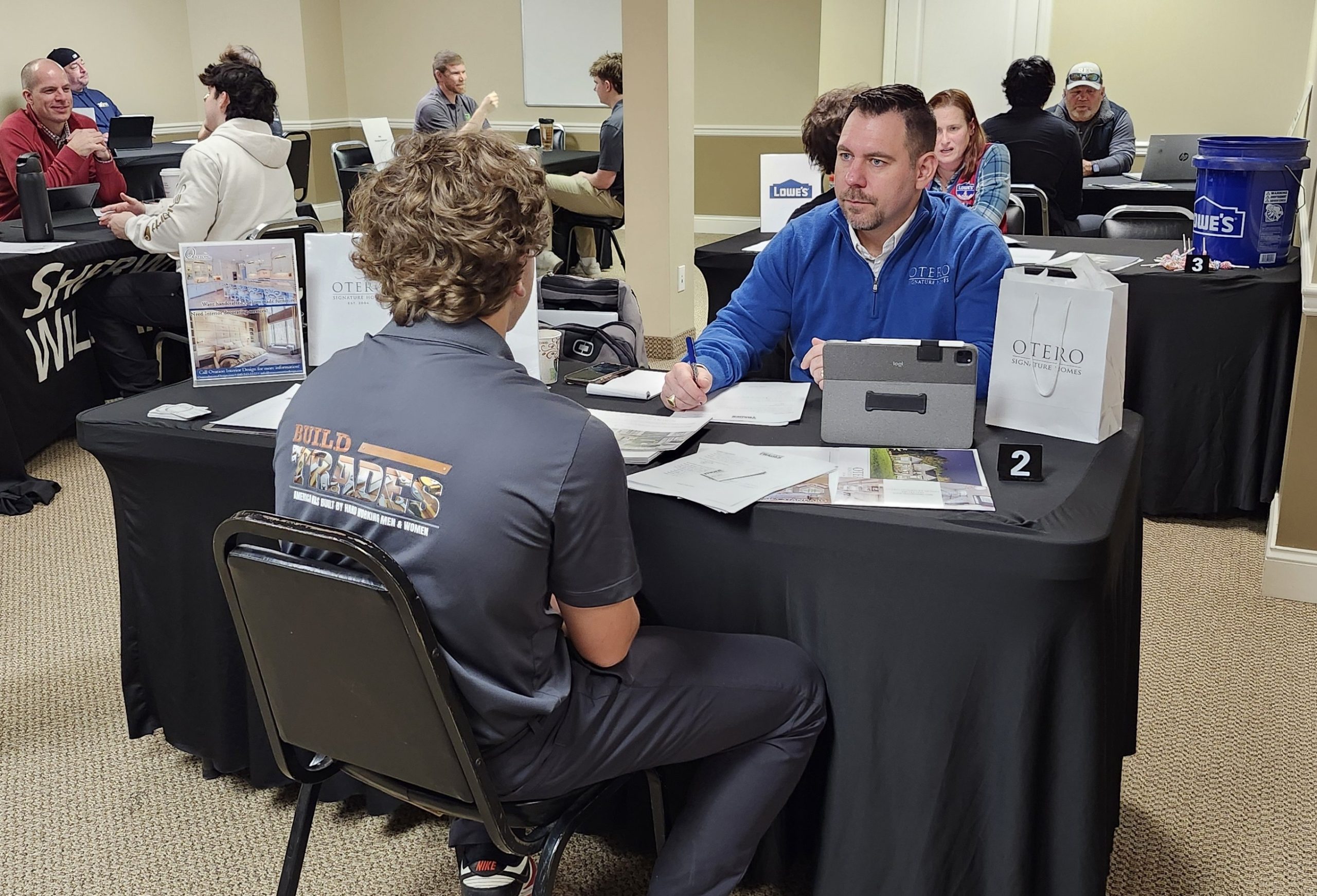 Two men sit across from each other at a trade-show table, discussing with papers and laptops nearby in a conference room setting.